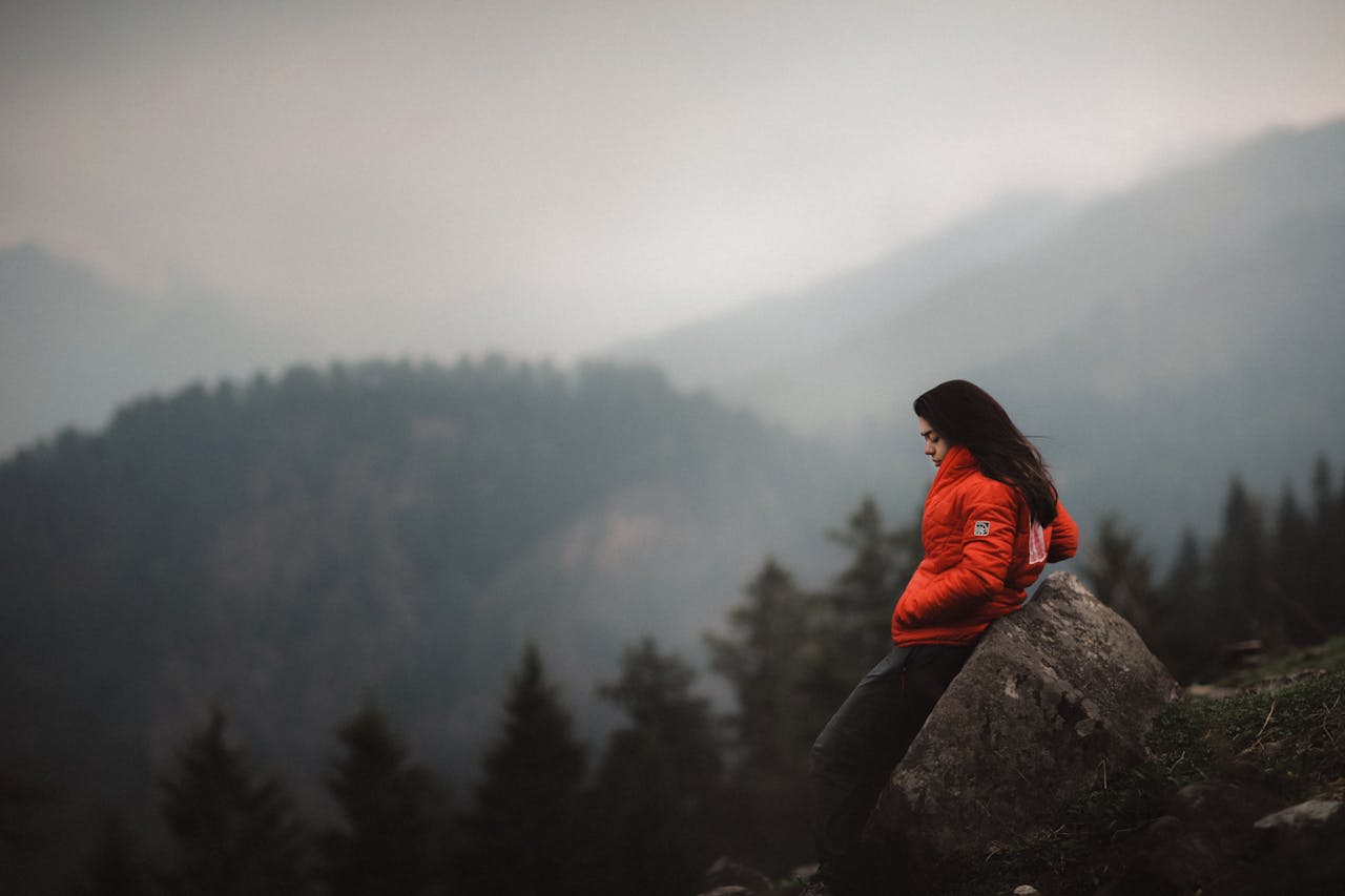 services-01 A woman in a red jacket leans against a boulder, enjoying the mountainous view in India.